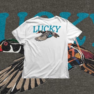 Flying duck PNG design with a colorful wood duck in side view, detailed wings, black ink linework, and large Lucky typography across the top. This digital artwork works well for duck lover shirts, nature themed sweatshirts, outdoor apparel, and printable creative products.