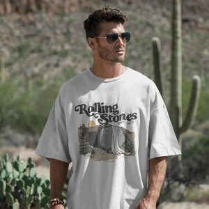 May include: A light gray t-shirt featuring the Rolling Stones logo and a desert-themed graphic. The graphic includes a tomb, sun, and desert plants. The shirt is worn by a person wearing sunglasses.