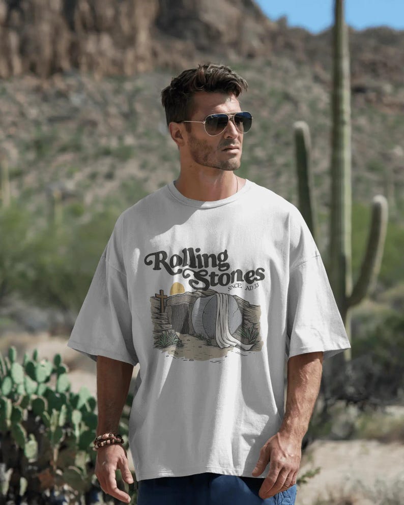 May include: A light gray t-shirt featuring the Rolling Stones logo and a desert-themed graphic. The graphic includes a tomb, sun, and desert plants. The shirt is worn by a person wearing sunglasses.