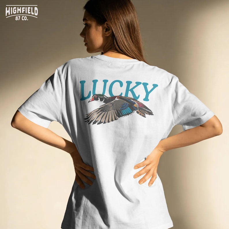 Bird lover PNG featuring a wood duck in flight with strong wing movement, bright eye detail, colorful feathers, and large Lucky text. The clean composition works well for t-shirts, sweatshirts, stickers, nature gifts, and outdoor digital design projects.