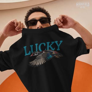 Lucky Duck shirt design showing a colorful flying duck with blue, brown, black, green, and red feather details under bold teal lettering. This PNG artwork is suitable for outdoor apparel, bird themed shirts, waterfowl designs, and nature inspired products.