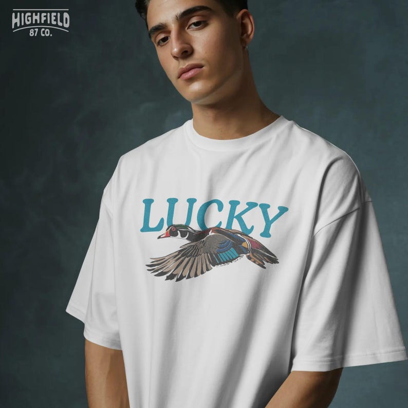 Wood duck shirt PNG with a flying waterfowl bird, colorful wing markings, red eye, black outlines, and bold Lucky typography. The artwork has a vintage outdoor feel for casual shirts, nature apparel, bird gifts, and printable digital products.