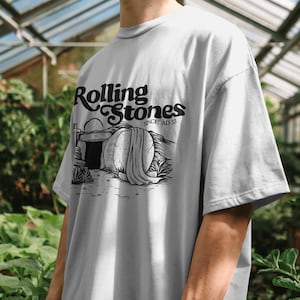 May include: A light gray t-shirt featuring the text "Rolling Stones SINCE AD 33" in black script. The graphic depicts a tomb with a draped stone and a sunrise. The shirt is a casual, oversized fit.