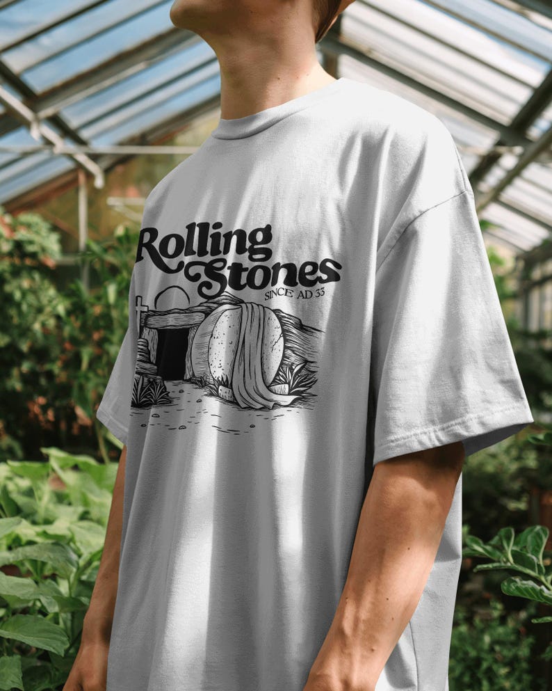 May include: A light gray t-shirt featuring the text "Rolling Stones SINCE AD 33" in black script. The graphic depicts a tomb with a draped stone and a sunrise. The shirt is a casual, oversized fit.