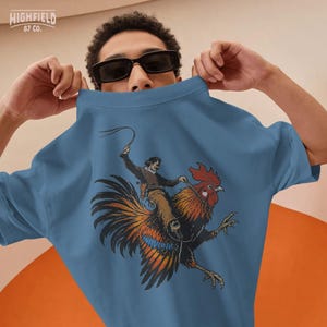 Rooster riding cowboy PNG artwork with a humorous western theme, showing a man riding a large colorful rooster in motion for novelty shirt designs.