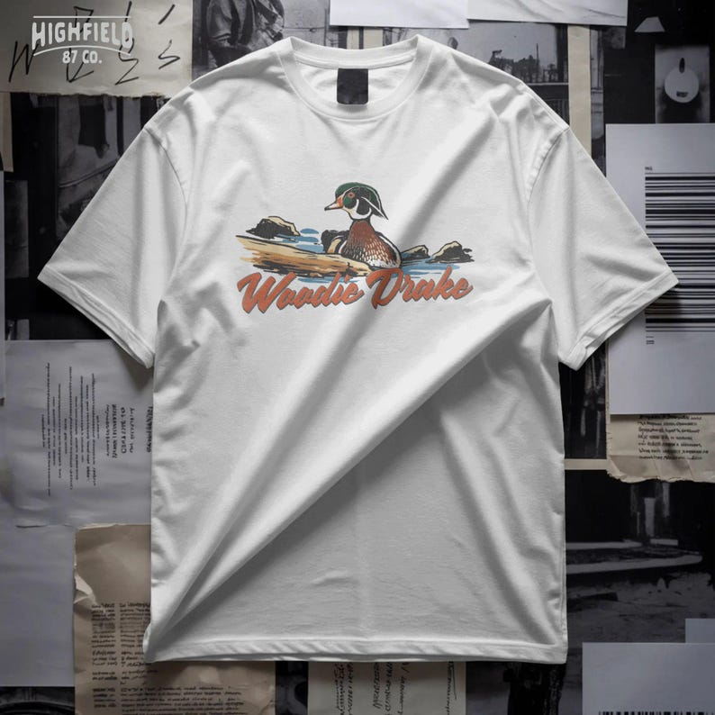 Hand drawn wood duck PNG with rocks and water details and Woodie Drake text, vintage duck hunting illustration
