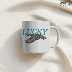 Vintage duck PNG illustration featuring a wood duck flying under teal Lucky text with bold black linework and flat natural colors. This design is suitable for nature shirts, bird watcher gifts, outdoor apparel, and printable PNG projects.