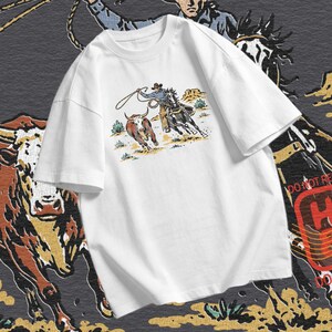 Cowboy roping PNG showing a rider, horse, rope, steer, desert plants, and western rodeo artwork for country apparel projects.