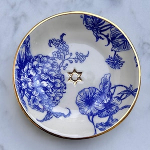Può includere: Un piattino rotondo in ceramica con base bianca e motivi floreali blu. Il bordo è rifinito con una bordatura color oro. Al centro del piatto si trova un piccolo emblema color oro a forma di Stella di David, che spicca sul disegno floreale.