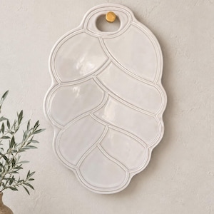 May include: A glossy, off-white ceramic serving platter shaped like a stylised pinecone or artichoke. The platter features a segmented, overlapping scale pattern and a circular cutout at the top, which is suspended by a small, round gold-coloured knob.