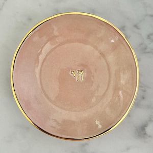 Puede incluir: Un plato redondo de cerámica rosa pálido con un acabado liso y brillante y un borde fino pintado en oro. Centrado en el medio del plato está el carácter hebreo Chai, representado en un acabado dorado metálico en relieve sobre el rosa.
