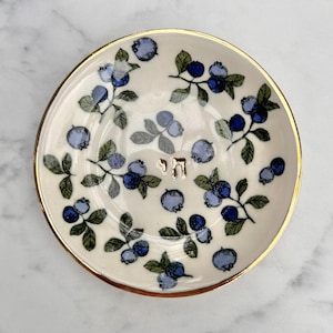 Può includere: Un piattino rotondo in ceramica con base color crema caratterizzato da un motivo ripetuto di mirtilli blu e foglie verdi. Il bordo è rifinito con un sottile filo metallico dorato. Al centro, le lettere ebraiche 'chai' sono in oro.