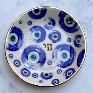 Puede incluir: Un plato pequeño redondo de cerámica con un patrón pintado a mano de motivos de ojo turco en azul y blanco. El centro del plato muestra la letra hebrea Chai en un acabado dorado metálico, y el borde está acentuado con un fino borde dorado.