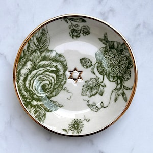 Puede incluir: Un plato pequeño redondo de cerámica con base color crema, que presenta un estampado botánico floral verde y un borde pintado en oro. En el centro del plato hay un pequeño símbolo en relieve de la Estrella de David de seis puntas en dorado.