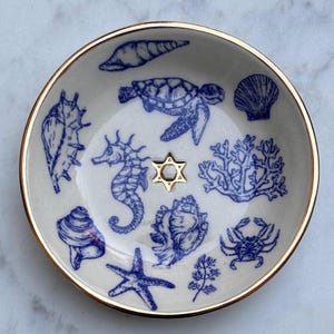 Peut inclure: Un vide-poche rond en céramique avec une base couleur crème et un bord doré métallique. L'intérieur présente des illustrations à l'encre bleue d'une tortue marine, d'un hippocampe, d'une étoile de mer, d'un crabe, de corail et de divers coquillages.