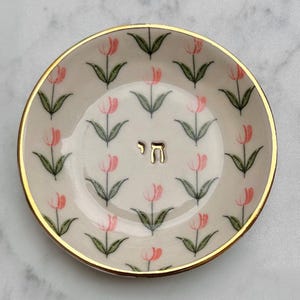 Op de afbeelding: Een rond keramieken schaaltje met een crèmekleurige basis en een herhalend patroon van roze tulpen met groene stelen en bladeren. De rand is afgewerkt met een goudkleurige boord en in het midden staat het Hebreeuwse teken Chai in goud.