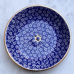 Puede incluir: Un cuenco redondo de cerámica con un fondo azul intenso cubierto por un patrón repetitivo de pequeñas flores de cerezo blancas, acabado con un borde dorado metálico y un emblema dorado de la Estrella de David en el centro.