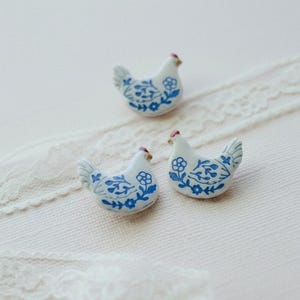 May include: Three small ceramic chicken-shaped ornaments featuring a white base with blue floral patterns and a tiny red accent on the comb. Each piece is shaped like a sitting hen with detailed blue painted leaves and flowers across the body.