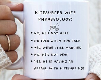 May include: White ceramic mug with black text that reads "Kitesurfer Wife Phraseology:" and a list of humorous phrases about a kitesurfer's wife. The mug is being held by a hand wearing gold rings.
