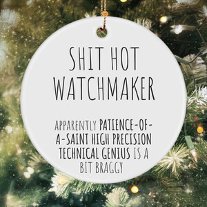 May include: White round Christmas ornament with black text that reads "Shit Hot Watchmaker. Apparently patience-of-a-saint high precision technical genius is a bit braggy."