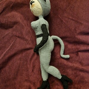 K&ouml;nnte beinhalten: Geh&auml;kelte Puppe im Katzenkost&uuml;m. Die Puppe ist &uuml;berwiegend grau mit schwarzen Akzenten, einschlie&szlig;lich Ohren, G&uuml;rtel und Stiefeln. Das Gesicht ist beige mit schwarzen Augen und einem kleinen L&auml;cheln. Die Puppe hat einen Schwanz und ist in einer Laufposition dargestellt.