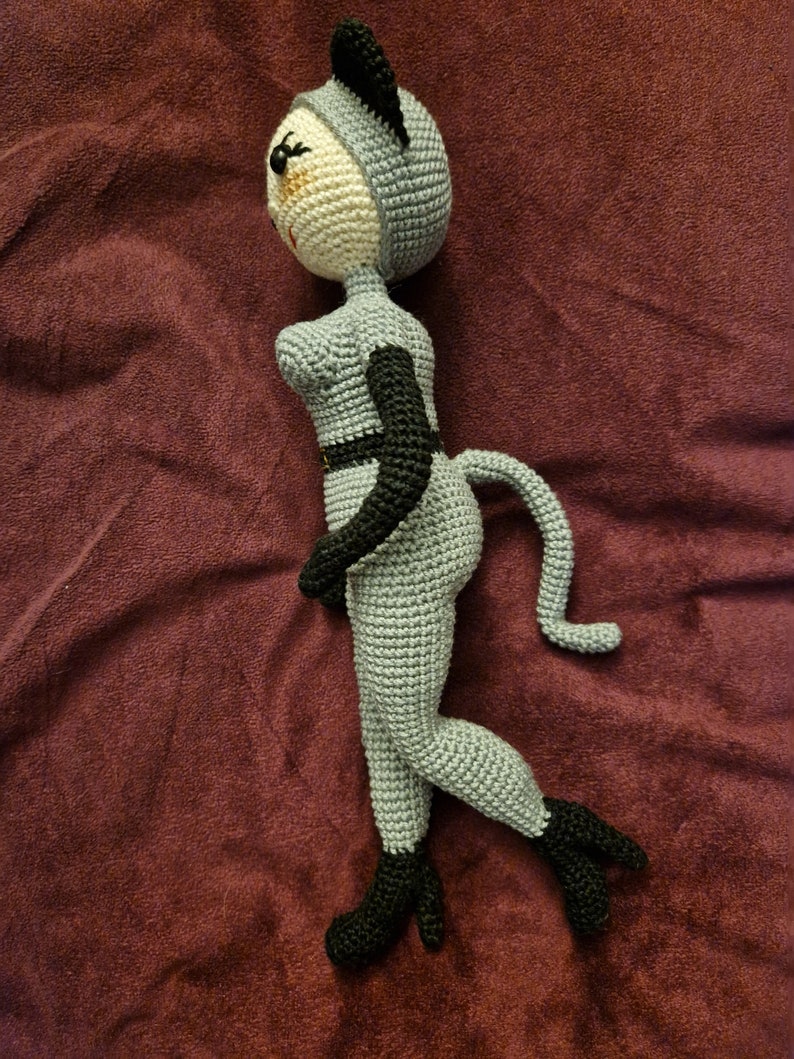 K&ouml;nnte beinhalten: Geh&auml;kelte Puppe im Katzenkost&uuml;m. Die Puppe ist &uuml;berwiegend grau mit schwarzen Akzenten, einschlie&szlig;lich Ohren, G&uuml;rtel und Stiefeln. Das Gesicht ist beige mit schwarzen Augen und einem kleinen L&auml;cheln. Die Puppe hat einen Schwanz und ist in einer Laufposition dargestellt.