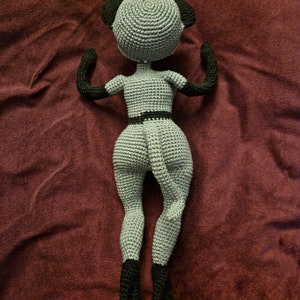 K&ouml;nnte beinhalten: Eine handgefertigte geh&auml;kelte Puppe in Grau und Schwarz. Die Puppe hat einen runden Kopf mit schwarzen Ohren, schwarzen Armen und schwarzen Stiefeln. Die Puppe tr&auml;gt einen schwarzen G&uuml;rtel. Die Puppe liegt auf einer weinroten Oberfl&auml;che.