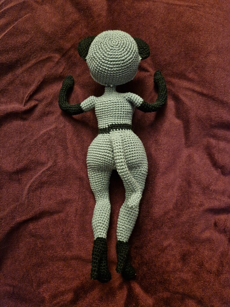 K&ouml;nnte beinhalten: Eine handgefertigte geh&auml;kelte Puppe in Grau und Schwarz. Die Puppe hat einen runden Kopf mit schwarzen Ohren, schwarzen Armen und schwarzen Stiefeln. Die Puppe tr&auml;gt einen schwarzen G&uuml;rtel. Die Puppe liegt auf einer weinroten Oberfl&auml;che.