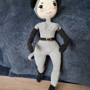 K&ouml;nnte beinhalten: Handgefertigte H&auml;kelpuppe in Grau und Schwarz. Die Puppe tr&auml;gt einen grauen Overall, schwarze Handschuhe und eine passende Kopfbedeckung mit schwarzen Ohren. Das Gesicht der Puppe hat rosige Wangen und ein rotes L&auml;cheln, mit einem schwarzen G&uuml;rtel.