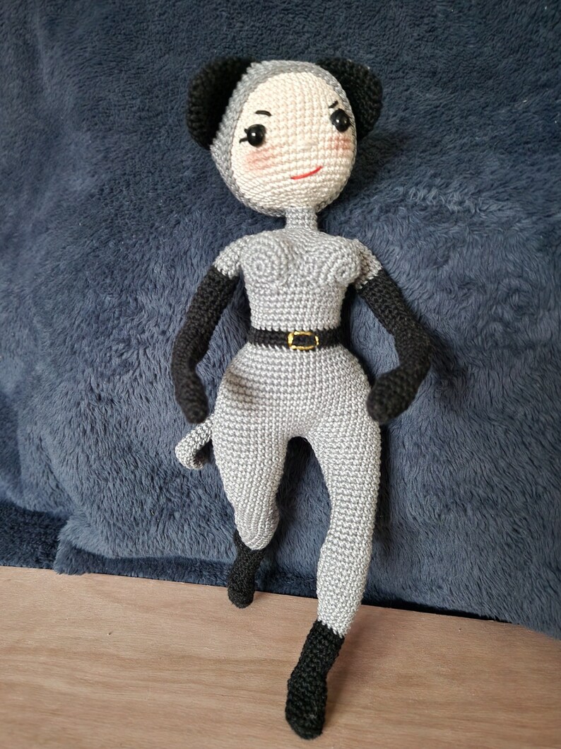 K&ouml;nnte beinhalten: Handgefertigte H&auml;kelpuppe in Grau und Schwarz. Die Puppe tr&auml;gt einen grauen Overall, schwarze Handschuhe und eine passende Kopfbedeckung mit schwarzen Ohren. Das Gesicht der Puppe hat rosige Wangen und ein rotes L&auml;cheln, mit einem schwarzen G&uuml;rtel.