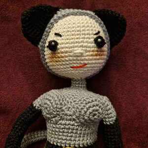 K&ouml;nnte beinhalten: Handgemachte H&auml;kelpuppe in Grau und Schwarz. Die Puppe hat ein hellbeige Gesicht mit schwarzen Augen, einem roten Mund und schwarzen Ohren. Die Puppe tr&auml;gt ein graues Kleid mit schwarzem G&uuml;rtel und goldener Schnalle.