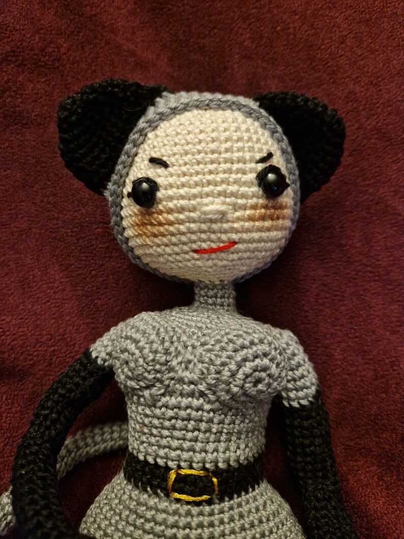 K&ouml;nnte beinhalten: Handgemachte H&auml;kelpuppe in Grau und Schwarz. Die Puppe hat ein hellbeige Gesicht mit schwarzen Augen, einem roten Mund und schwarzen Ohren. Die Puppe tr&auml;gt ein graues Kleid mit schwarzem G&uuml;rtel und goldener Schnalle.