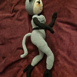 K&ouml;nnte beinhalten: Handgefertigte H&auml;kelpuppe in Grau und Schwarz. Die Puppe hat ein katzenartiges Aussehen mit einem grauen K&ouml;rper, schwarzen Ohren, Armen und F&uuml;&szlig;en. Die Puppe ist dynamisch posiert, mit einem Arm erhoben und dem anderen ausgestreckt.