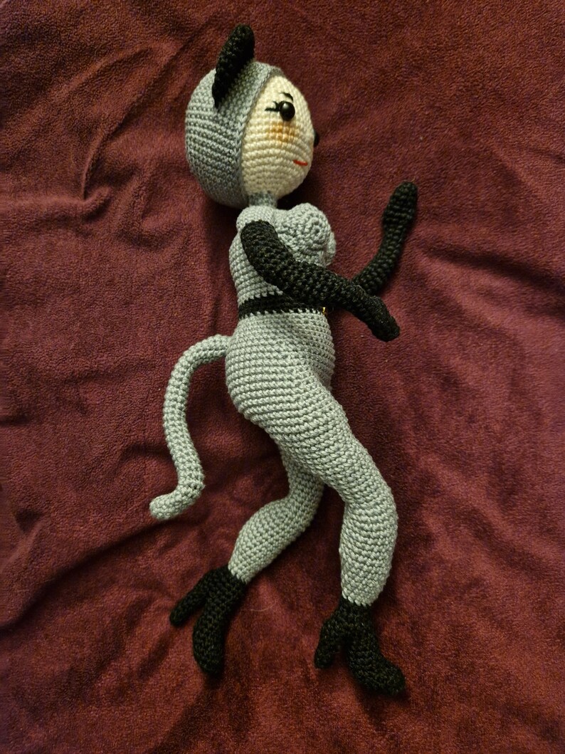 K&ouml;nnte beinhalten: Handgefertigte H&auml;kelpuppe in Grau und Schwarz. Die Puppe hat ein katzenartiges Aussehen mit einem grauen K&ouml;rper, schwarzen Ohren, Armen und F&uuml;&szlig;en. Die Puppe ist dynamisch posiert, mit einem Arm erhoben und dem anderen ausgestreckt.