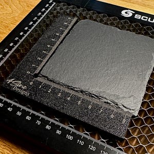 May include: A dark gray, rectangular slate coaster with a textured surface. A black ruler with white markings and the text "ASine Designs" is placed on the coaster. The ruler measures up to 6 inches.