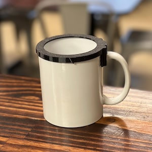 May include: A white ceramic mug with a black, circular lid. The lid appears to be made of a textured material and fits snugly around the mug's rim. The mug has a curved handle and sits on a wooden surface.