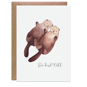 Op de afbeelding: Een wenskaart met een aquarel illustratie van twee otters die elkaar omhelzen. De otters zijn afgebeeld in bruin- en beigetinten tegen een witte achtergrond. De kaart zit in een bruine envelop en de tekst "Du bist toll" is geschreven in een scriptlettertype.