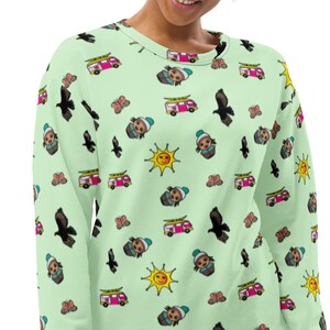May include: A light green sweatshirt with a repeating pattern of cartoon illustrations. The illustrations include a sun, a bird, a van, and a person wearing a hat.