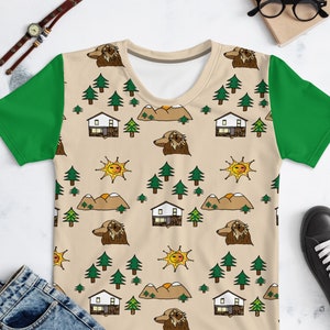 Puede incluir: Una camiseta para mujer beige y verde con un patrón repetitivo de casas, montañas, soles y árboles. El patrón es de estilo de dibujos animados.