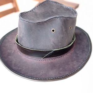 May include: A brown leather hat with a wide brim and a green leather band. The hat has two brass eyelets on the crown.