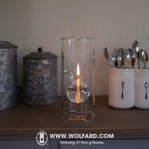 May include: A clear glass oil lamp with a flame burning inside. The lamp is sitting on a wooden surface with two metal canisters and a white ceramic utensil holder.