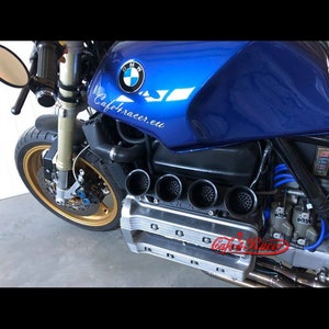 Può includere: Una moto blu con un logo BMW sul serbatoio. La moto ha un sistema di scarico personalizzato con quattro grossi tubi. La moto è appoggiata su una superficie grigia. Il testo "Cafe4racer.eu" è stampato sul serbatoio.