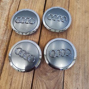 Könnte beinhalten: Vier silber-graue Audi Radnabenabdeckungen. Jede Kappe zeigt das Audi-Logo, ein Satz von vier ineinandergreifenden Ringen, in polierter Silberausführung auf grauem Hintergrund. Die Kappen sind kreisförmig mit einem polierten Silberrand.