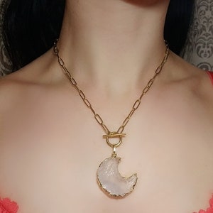 Op de afbeelding: Een gouden ketting met een halve maan hanger gemaakt van witte agaat. De hanger is ingelegd met goud en heeft een ruwe, natuurlijke textuur.