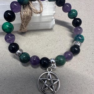 May include: A bracelet made of black, purple, and green gemstone beads. A silver-coloured pentagram charm is attached. The bracelet is strung on a cord.