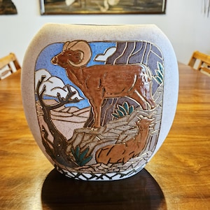 May include: A beige ceramic vase with a detailed relief of a ram and a resting animal against a blue and tan background. The vase has a rounded shape and sits on a wooden table. The artwork features brown, green, and tan colors.