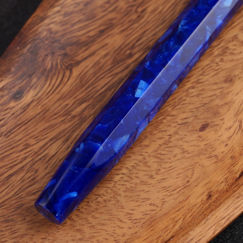 May include: Close-up of a faceted, deep blue pen. The pen's body has a marbled appearance with lighter blue swirls, creating a unique pattern. The pen is set against a wooden surface.