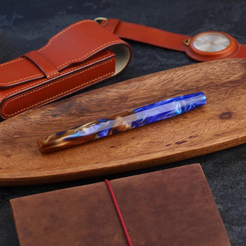 May include: A fountain pen with a marbled blue, white, and brown resin barrel. The pen rests on a wooden tray alongside a brown leather pen case, a brown leather notebook with a red elastic band, and a brown leather wristwatch with a round face.