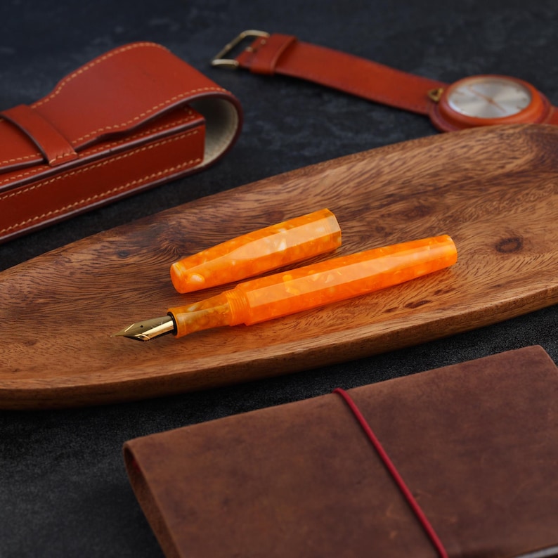 May include: An orange and cream-colored fountain pen with a gold nib rests on a wooden tray. A leather watch with a brown strap and a leather pen case are in the background. A brown leather journal is in the foreground.