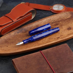 May include: A blue fountain pen with a silver nib and a matching cap rests on a wooden tray. The pen has a marbled design. A leather pen case, a watch, and a leather-bound notebook are also visible.